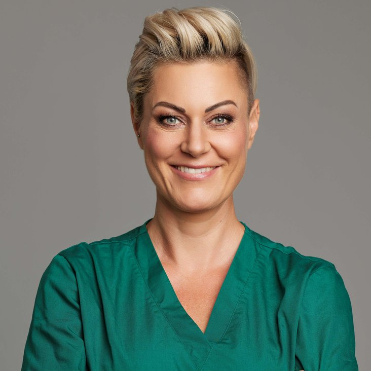Doc Caro mit einem grünen Arztkittel. Sie hat kurze blonden Haare und tätowierte Arme, die vor dem Körper verschränkt sind.