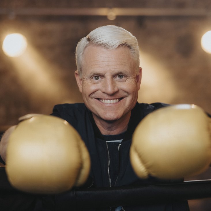 Der blonde Entertainer Guido Cantz lacht in die Kamera. Er hat zwei goldene Boxhandschuhe an, die er in die Kamera streckt.