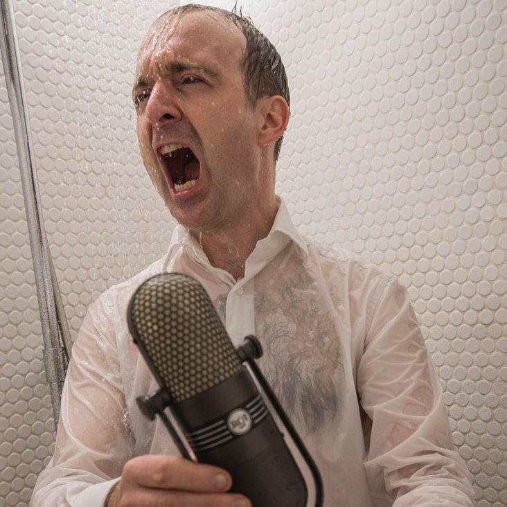 Patrick Bopp singt aus voller Kehle mit nasser Kleidung in einer Dusche, er hält ein Mikrofon in der Hand