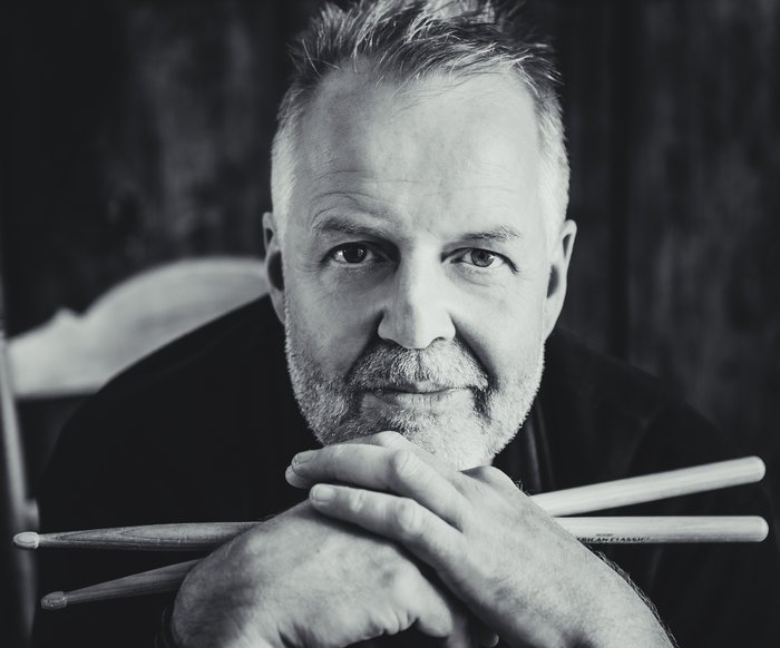 Portrait in schwarz weiß von Wolfgang Haffner. Er stützt seinen Kopf auf seine Hände, in denen er zwei Drumsticks hält.