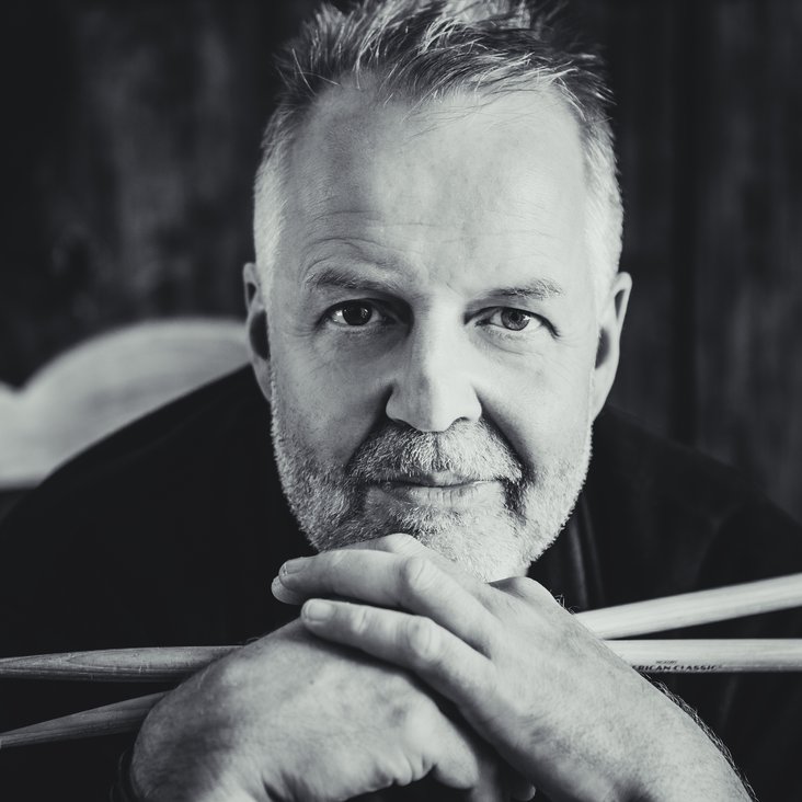 Portrait in schwarz weiß von Wolfgang Haffner. Er stützt seinen Kopf auf seine Hände, in denen er zwei Drumsticks hält.