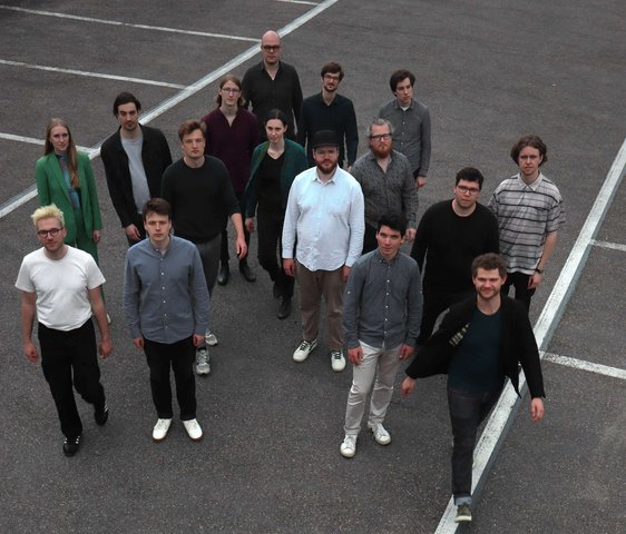 Gruppenfoto der Samuel Restle Bigband. Die Musiker:innen stehen auf einem leeren Parkplatz und werden von oben oder aus erhöhter Perspektive fotografiert.