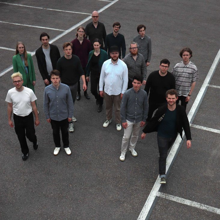 Gruppenfoto der Samuel Restle Bigband. Die Musiker:innen stehen auf einem leeren Parkplatz und werden von oben oder aus erhöhter Perspektive fotografiert.