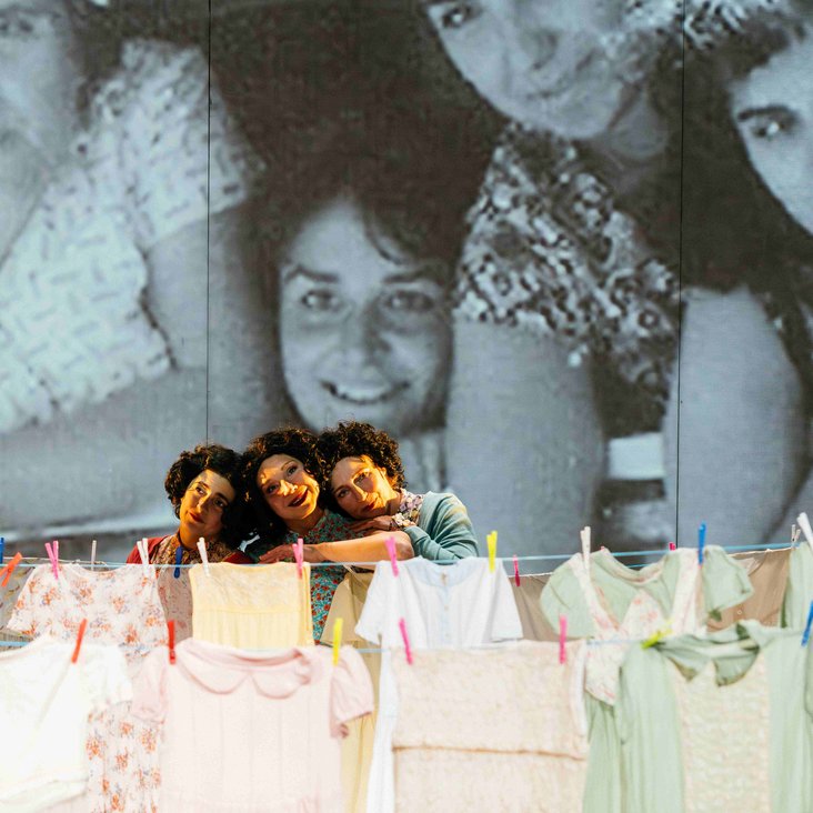 Drei Frauen auf der Bühne blicken über eine Wäscheleine in die Ferne, im Hintergrund eine Bühnenprojektion eines schwarz-weißen Familienfotos