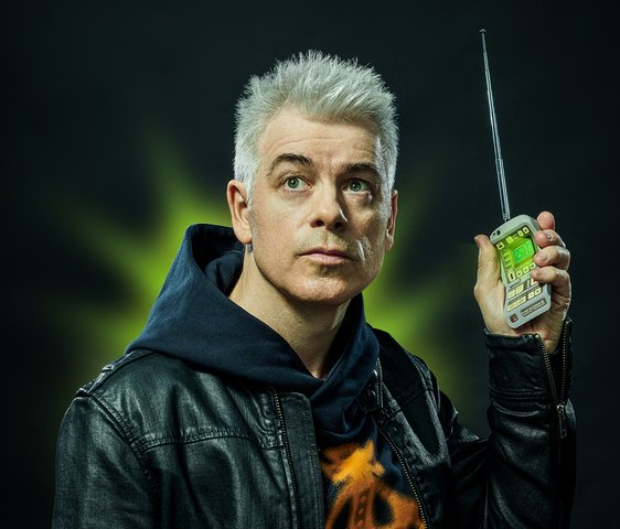 Michael Mittermeier trägt einen Kapuzenpullover und darüber eine Lederjacke. Er blickt in die Ferne und hält in seiner linken Hand ein technisches Gerät in Handygröße (Tricorder) nach oben. Es leuchtet grün, auch im Hintergrund ist grünes Licht zu sehen.