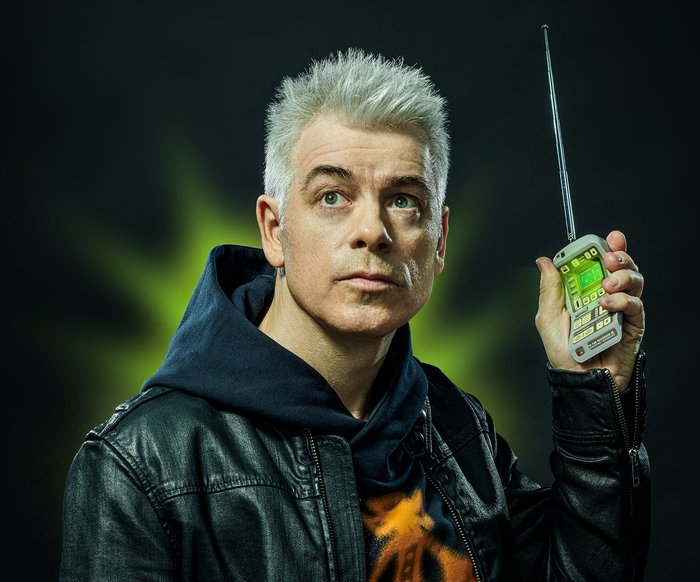 Michael Mittermeier trägt einen Kapuzenpullover und darüber eine Lederjacke. Er blickt in die Ferne und hält in seiner linken Hand ein technisches Gerät in Handygröße (Tricorder) nach oben. Es leuchtet grün, auch im Hintergrund ist grünes Licht zu sehen.