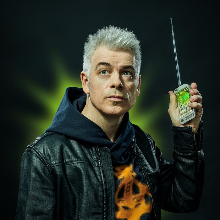 Michael Mittermeier trägt einen Kapuzenpullover und darüber eine Lederjacke. Er blickt in die Ferne und hält in seiner linken Hand ein technisches Gerät in Handygröße (Tricorder) nach oben. Es leuchtet grün, auch im Hintergrund ist grünes Licht zu sehen.