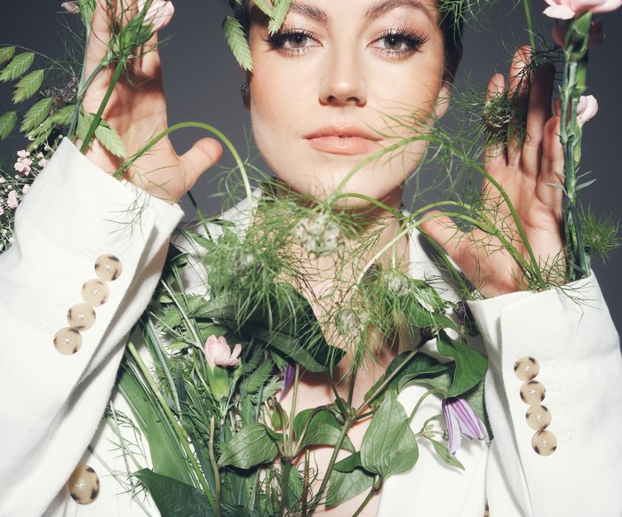 Portraitfoto von Tahnee in einem weißen Blazer mit verschiedenen Blüten und Blumen um sie und an ihr
