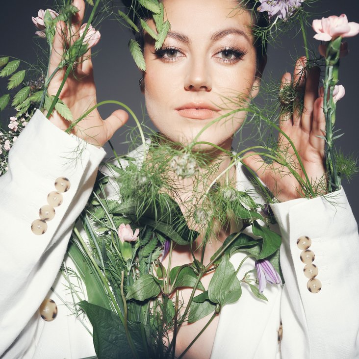 Portraitfoto von Tahnee in einem weißen Blazer mit verschiedenen Blüten und Blumen um sie und an ihr
