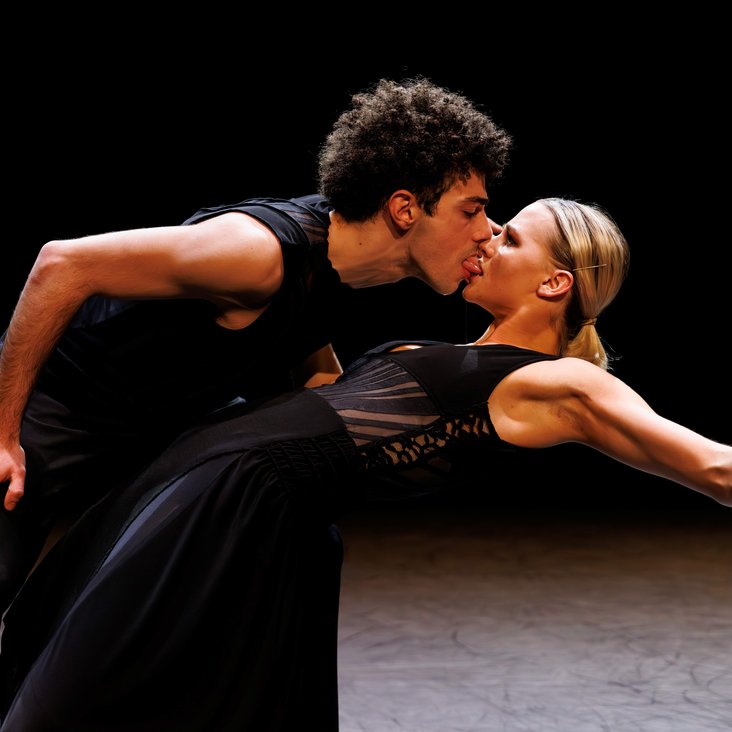 Tänzer:innen in Kostüm auf der Bühne, der Boden ist schwarz und die Bühne ist professionell ausgeleuchtet.