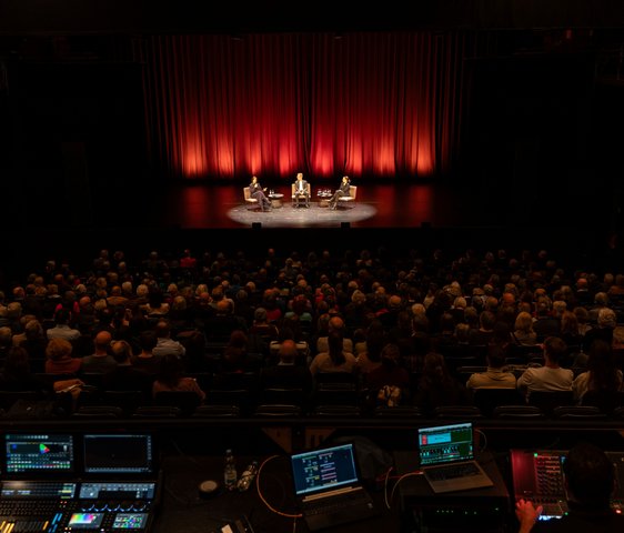 Ein Theatersaal, in den Rängen sitzen Menschen, im Hintergrund ist eine Gesprächssituation auf der Bühne. Im Vordergrund ist das Technikpult zu sehen.