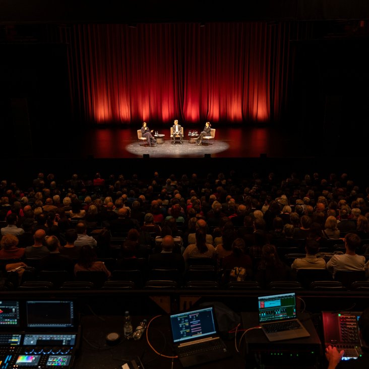 Ein Theatersaal, in den Rängen sitzen Menschen, im Hintergrund ist eine Gesprächssituation auf der Bühne. Im Vordergrund ist das Technikpult zu sehen.