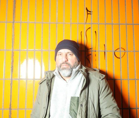 Serdar Somuncu steht vor einer gelb gefliesten Wand. Er trägt einen grünen Parka und eine graue Mütze. Er hat die Hände in die Taschen gesteckt und blickt ernst in die Kamera.