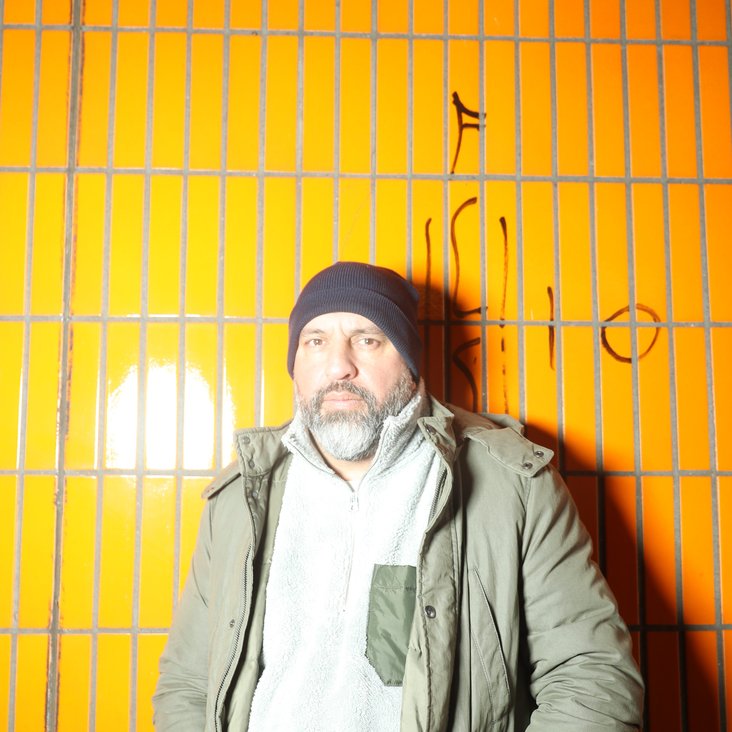 Serdar Somuncu steht vor einer gelb gefliesten Wand. Er trägt einen grünen Parka und eine graue Mütze. Er hat die Hände in die Taschen gesteckt und blickt ernst in die Kamera.