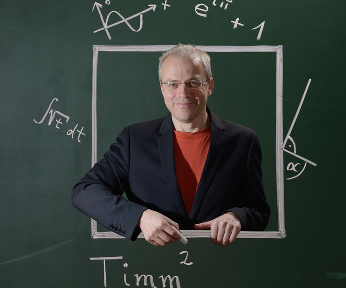 Timm Sigg steht vor einem Layout einer Kreidetafel mit Beschriftungen mathematischer Formeln