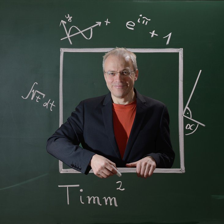 Timm Sigg steht vor einem Layout einer Kreidetafel mit Beschriftungen mathematischer Formeln