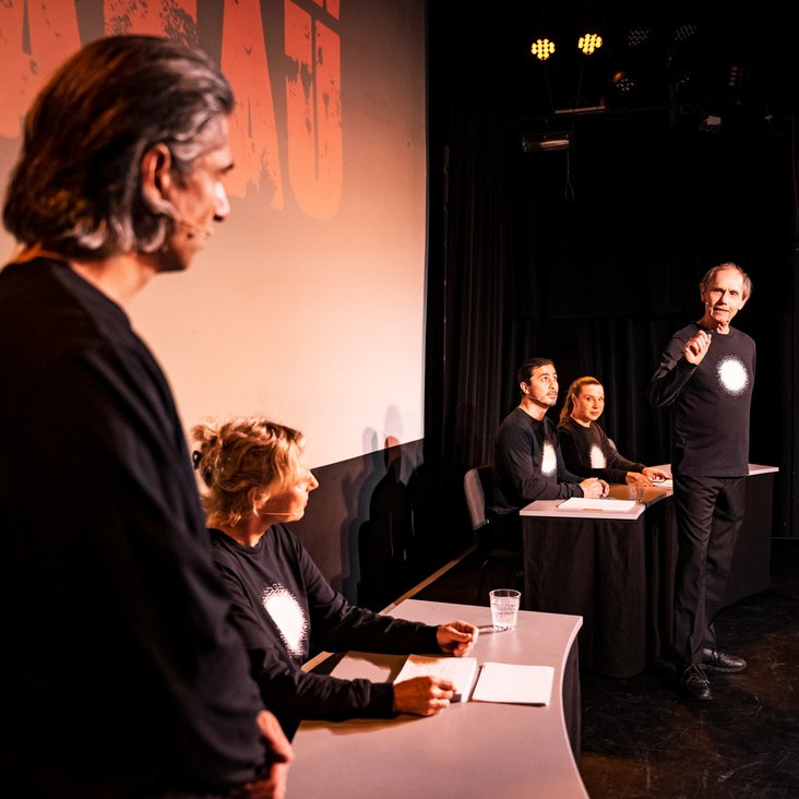 Die Schauspieler:innen von "And now Hanau" auf der Bühne. Sie tragen alle schwarze Kleidung mit einem vollflächig weißen Kreis auf der Brust.