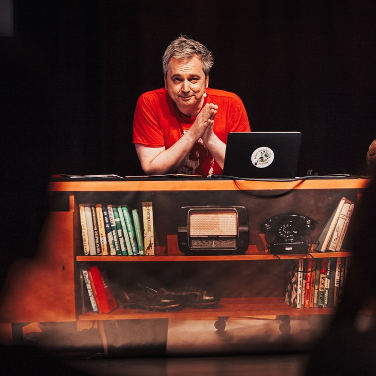 Arnd Zeigler sitzt auf einer Bühne an einem Schreibtisch. Vor im steht ein Laptop. Er trägt ein rotes T-Shirt und blickt in die Kamera.