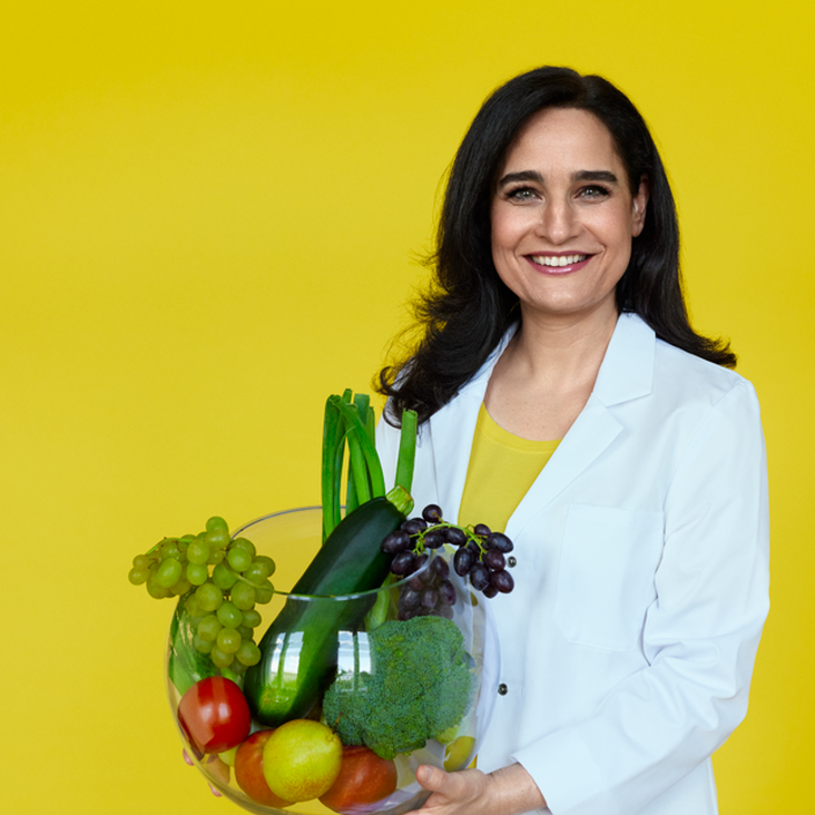Eine dunkelhaarige Frau steht in einem weißen Blazer vor einer gelben Wand. Sie hält eine große Glaskugel voller Obst und Gemüse.