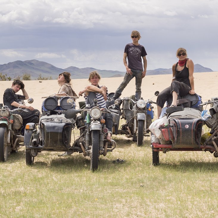 Fünf Personen stehen und sitzen auf ihren Motorrädern, die voller Gepäck sind auf einer Wiese mit Bergen im Hintergrund