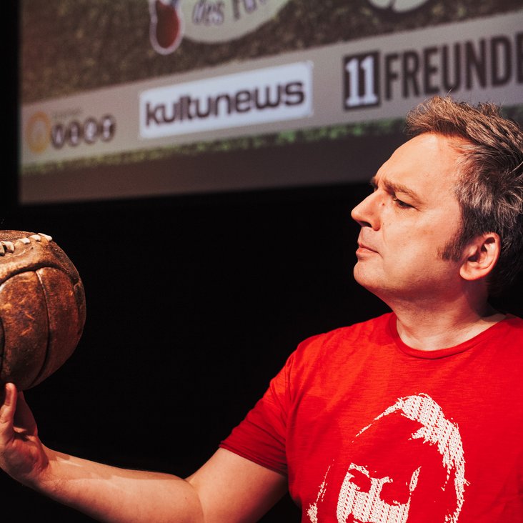 Arnd Zeigler steht in einem roten T-Shirt auf einer Bühne. Im Hintergrund ist eine Leinwand zu sehen. Arnd hat einen alten Lederfußball in der Hand und betrachtet ihn.
