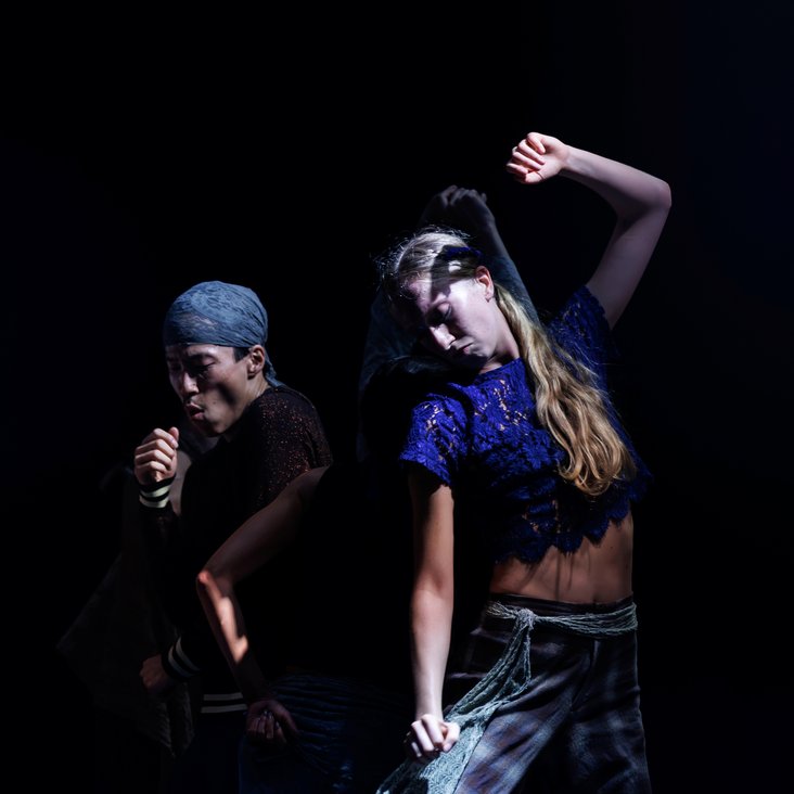 Die Tänzer:innen von Gauthier Dance in verschiedenfarbigen Kostümen auf der Bühne. Die Bühne ist auch dunkel und die Beleuchtung ist atmosphärisch.