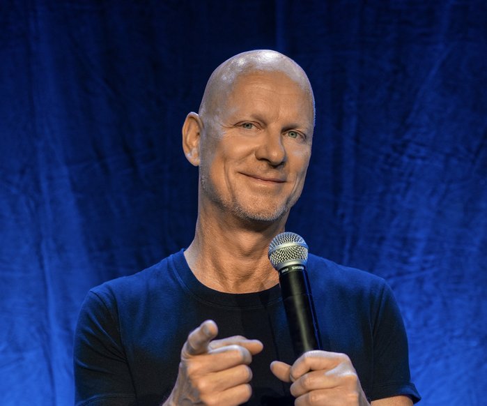 Rüdiger Hoffmann live auf der Bühne. Er trägt ein schwarzes T-Shirt und hat ein Mikrofon in der Hand. Er zeigt mit dem Zeigefinger in Richtung der Kamera und grinst.