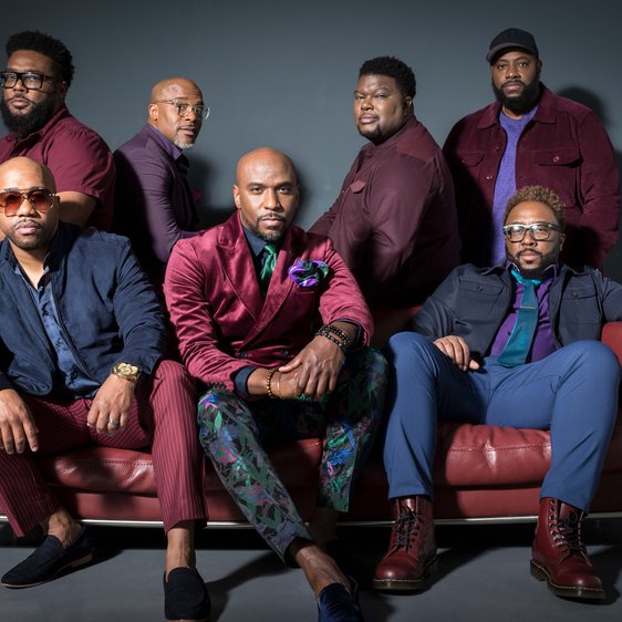 Die 7 Mitglieder von Naturally 7 sitzen auf und stehen um ein Sofa