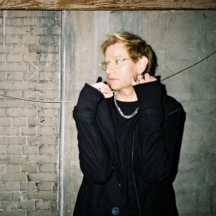 Porträt einer Person in dunkler Kleidung vor Backsteinwand, Hände am Kragen, seitlicher Blick.