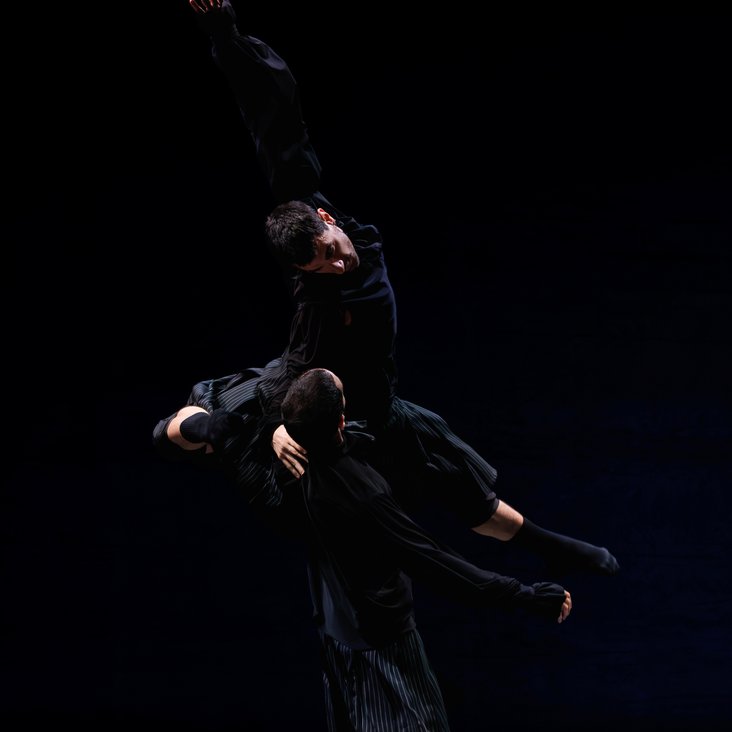 Die Tänzer:innen von Gauthier Dance in schwarzen Kostümen auf der Bühne. Die Bühne ist auch dunkel und es gibt wenig Licht.