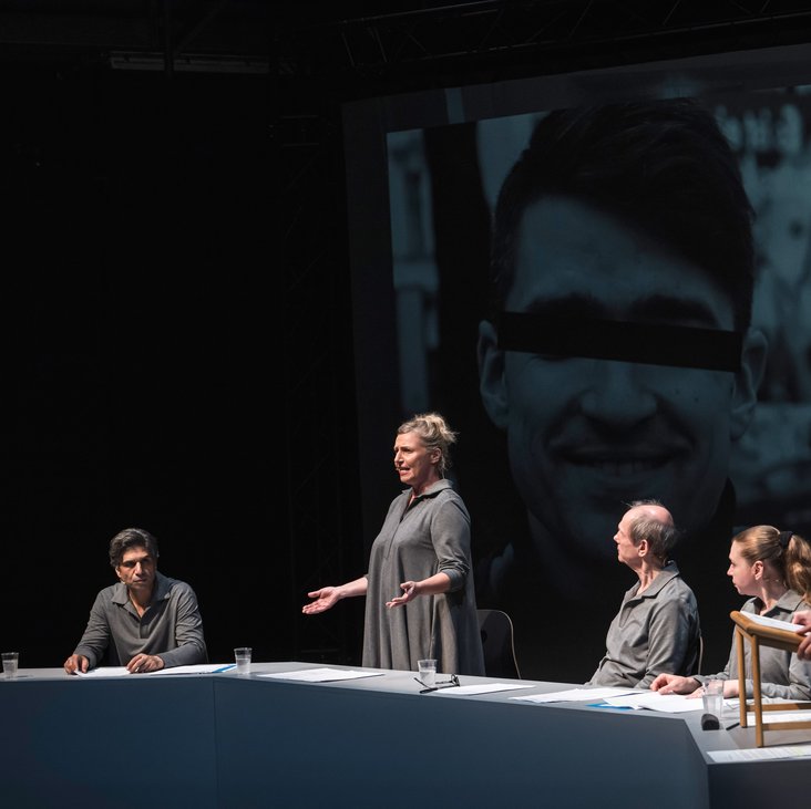 Szenenfoto, es zeigt fünf Schauspieler:innen sitzend und stehend in grauer Kleidung auf der Bühne. Im Hintergrund das schwarz-weiß Foto einer Person, die Augen sind zensiert.