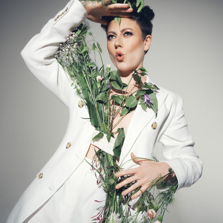 Portraitfoto von Tahnee in einem weißen Zweiteiler mit verschiedenen Blüten und Blumen um sie und an ihr