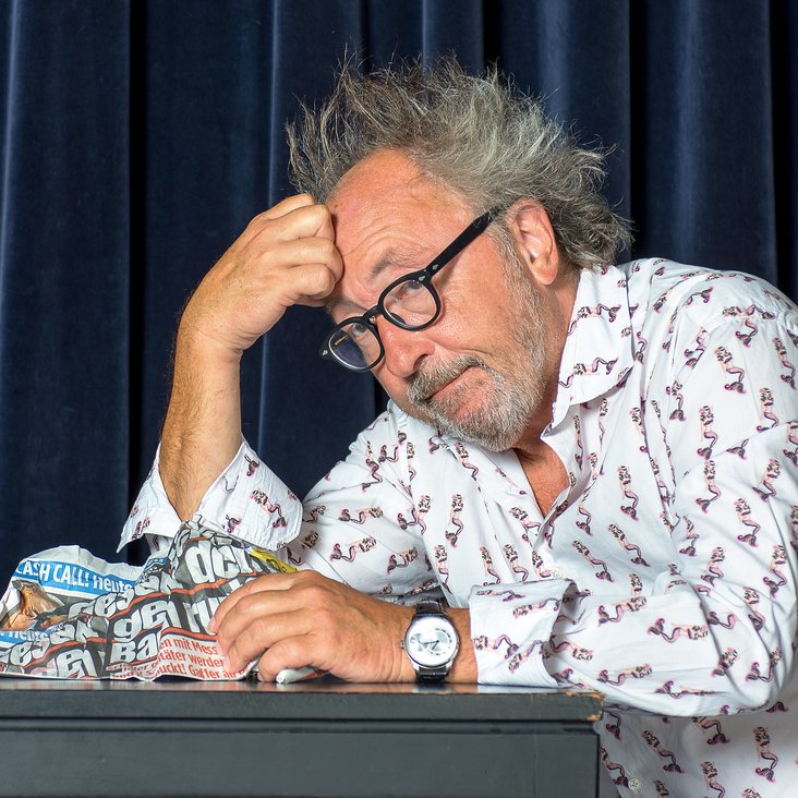 Portraitfoto Urban Priol. Er sitzt, den Kopf aufgestützt, mit einer Tageszeitung an einem Tisch auf der Bühne.
