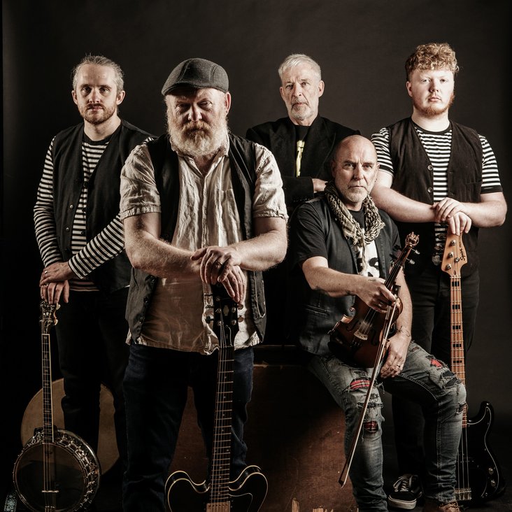 Die Musiker von The Outcast Crew stehen und sitzen mit ihren Instrumenten in der Gruppe und blicken in die Kamera