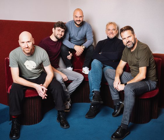 Die Bandmitglieder sitzen auf roten Polstermöbeln und schauen in die Kamera