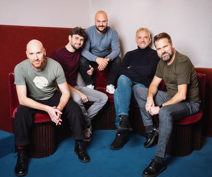 Die Bandmitglieder sitzen auf roten Polstermöbeln und schauen in die Kamera