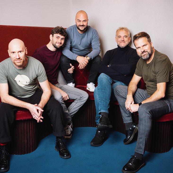 Die Bandmitglieder sitzen auf roten Polstermöbeln und schauen in die Kamera