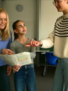 Drei Personen stehen im Klassenraum, betrachten Blatt und zeigen darauf.