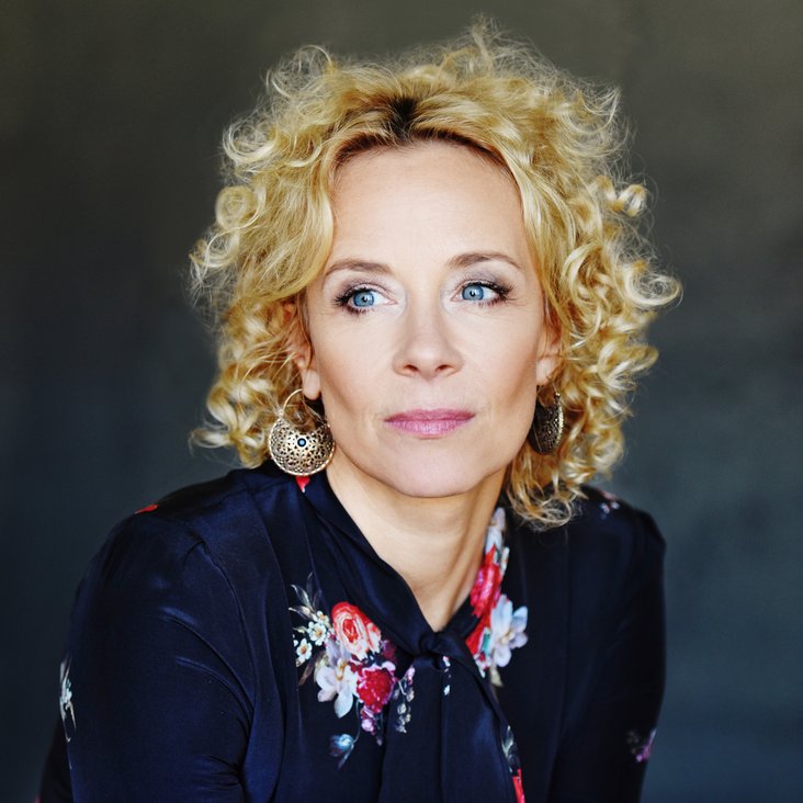 Katja Riemann mit lockigem, blondem Haar und einer schwarzen Bluse mit Muster