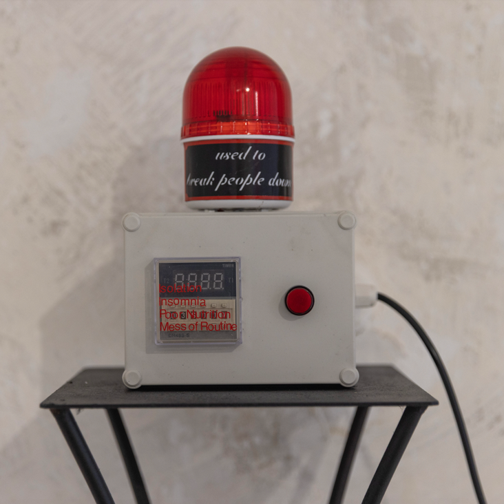 Das Foto zeigt ein technisches Gerät, das ein quadratischer grauer Kasten ist mit einer roten Alarmleuchte darauf. Es hat einen roten Knopf vorne und ein Kabel geht nach rechts weg. Auf der Leuchte steht: used to break people down. Auf dem grauen Kasten steht: Isolation, Insomnia, Poor Nutrition, Mess of Routine.