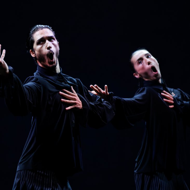 Die Tänzer:innen von Gauthier Dance in schwarzen Kostümen auf der Bühne. Die Bühne ist auch dunkel und es gibt wenig Licht.