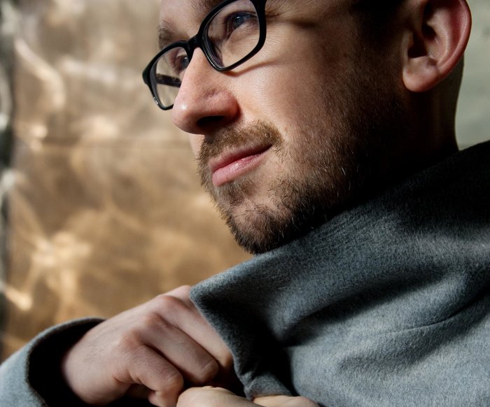 Der Künstler ist im PRofil zu sehen und trägt einen grauen Mantel und eine schwarze Brille.