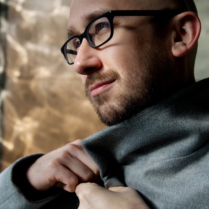 Der Künstler ist im PRofil zu sehen und trägt einen grauen Mantel und eine schwarze Brille.