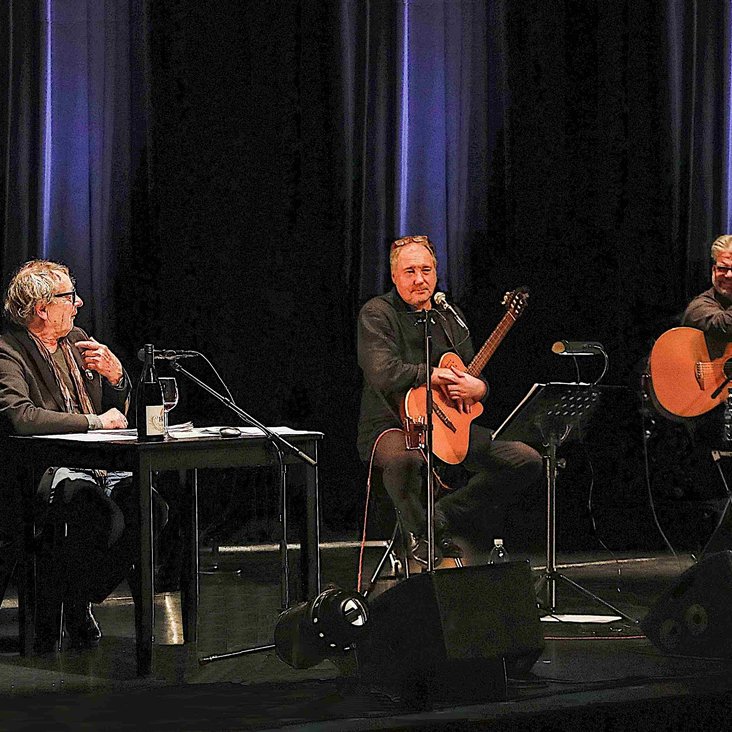 Die drei Künstler sitzen auf der Bühne an einem Tisch, bzw. auf Hockern. Die zwei Künstler auf den Hockern haben Gitarren in der Hand.