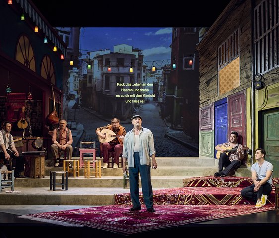 Ein Mann steht in der Mitte einer Bühne, hinter ihm Verteilt sitzt eine Band. Die Bühne zeigt eine Projektion Istanbuls.