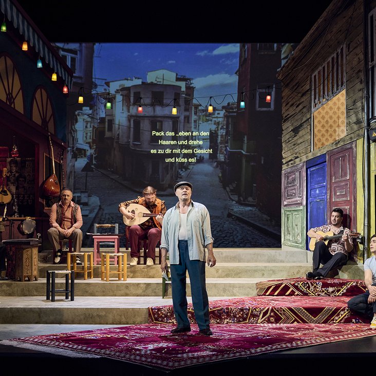 Ein Mann steht in der Mitte einer Bühne, hinter ihm Verteilt sitzt eine Band. Die Bühne zeigt eine Projektion Istanbuls.