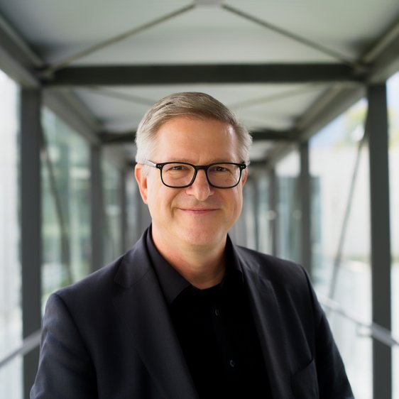 Person mit Brille und dunklem Sakko steht mittig in verglastem Gang.